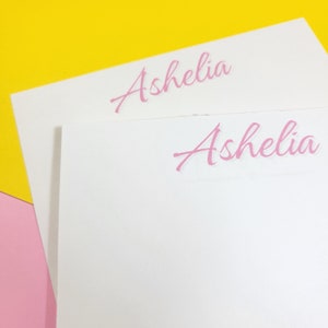 Op de afbeelding: Twee witte notitiekaartjes met de naam "Ashelia" in roze cursief op elk kaartje.