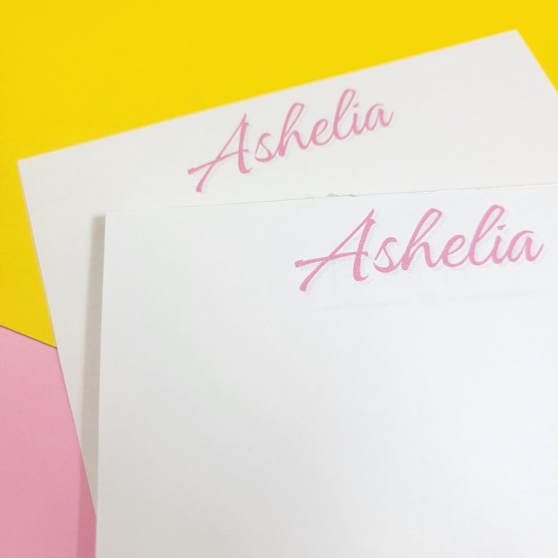 Op de afbeelding: Twee witte notitiekaartjes met de naam "Ashelia" in roze cursief op elk kaartje.