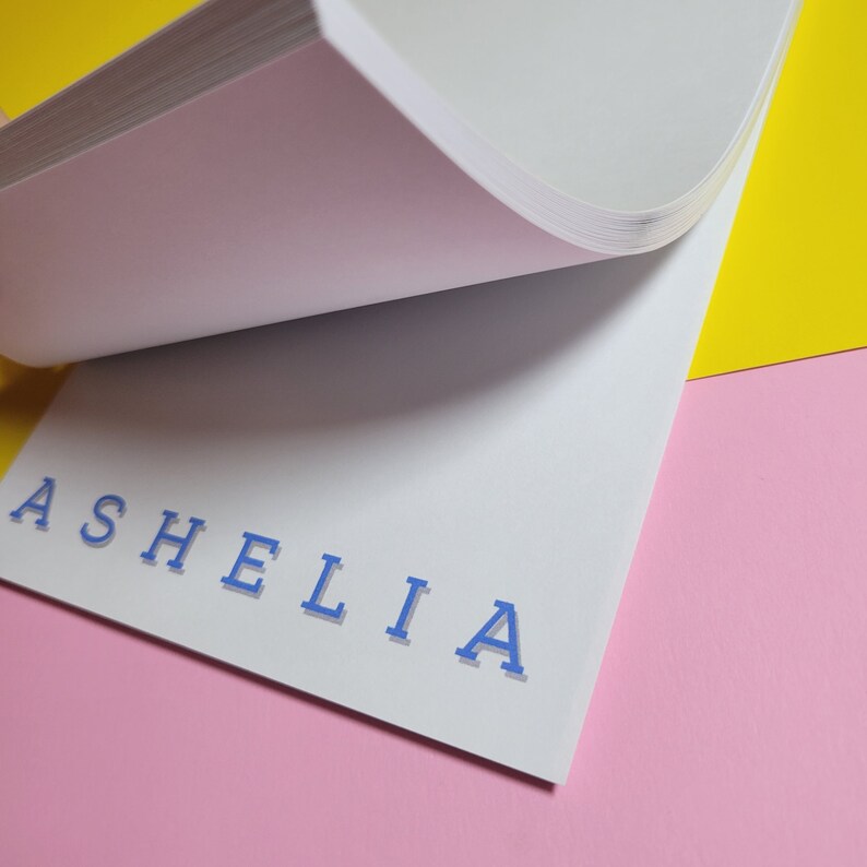 May include: A white notepad with the name "ASHELIA" written in blue letters on a pink and yellow background.