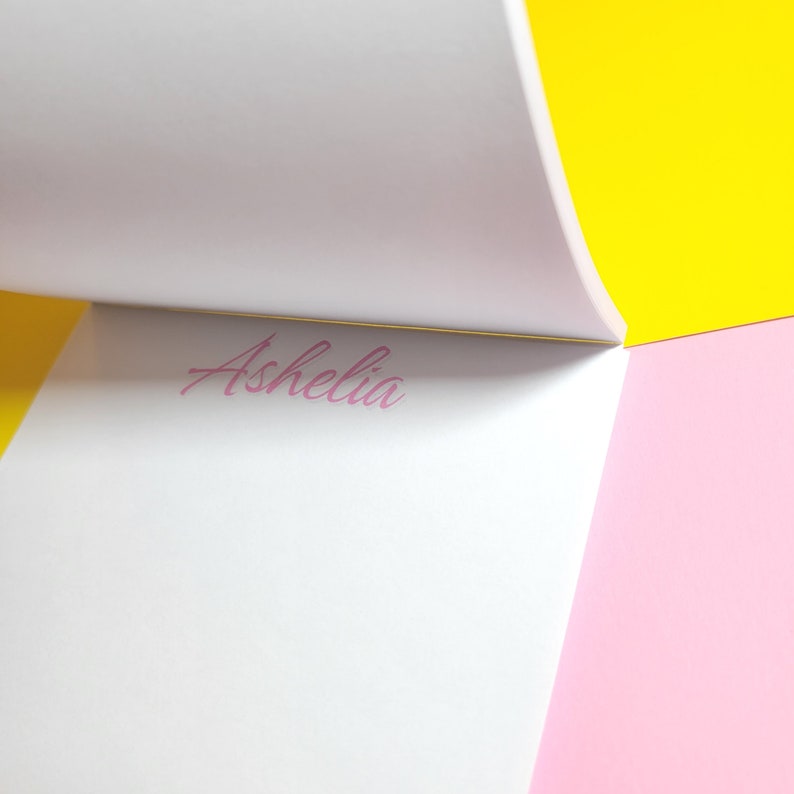 Op de afbeelding: Een wit notitieboekje met de naam "Ashelia" in roze cursief op de bovenste pagina. Het notitieboekje is open en de pagina's zijn omgeslagen, waardoor een gele achtergrond links en een roze achtergrond rechts zichtbaar worden.