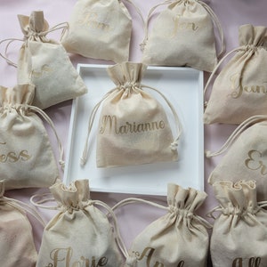 Puede incluir: Una colección de 10 pequeñas bolsas de tela beige con cordón y letras doradas. Cada bolsa tiene un nombre diferente escrito en ella, incluyendo Jess, Pam, Jon, Marianne, Janti, Ellie, Jo, Florie, Allison y Andy.