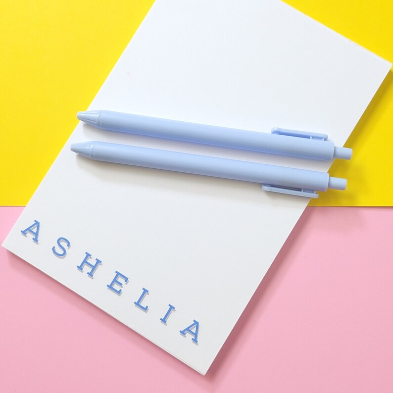 May include: A white notepad with the name "ASHELIA" printed in blue on a pink background. Two light blue pens are on top of the notepad.