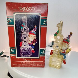Könnte beinhalten: Ein transparenter Weihnachtsbaumschmuck aus Glas mit einem kleinen Elf, der ein Stück Holz hält. Der Schmuck ist in Form der Zahl "1998" gestaltet und hat eine glitzernde Oberfläche. Die Schachtel trägt die Aufschrift "Enesco" und "Carver" sowie "Dritte Ausgabe der Wee Tree Trimmers Dated Series".