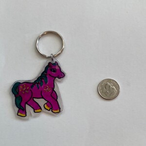 Puede incluir: Un llavero de plástico rosa y verde con un diseño de caballo. El llavero tiene un anillo de llave plateado.