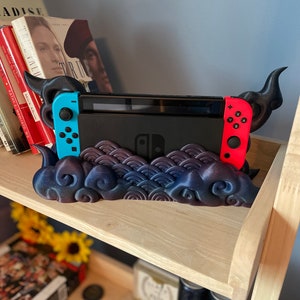 May include: A 3D printed stand for a Nintendo Switch console. The stand is black and blue with a galaxy design and features two red and black horns on either side. The console is sitting in the stand.
