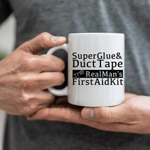 May include: White ceramic mug with the text "Super Glue & Duct Tape The Real Man's First Aid Kit" in black. The mug has a standard handle and is held by a person.