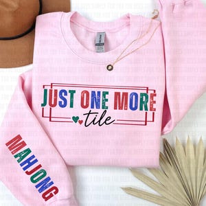 May include: A light pink sweatshirt with the text "JUST ONE MORE tile" in colorful letters. The sleeve reads "MAHJONG" in a matching color scheme. A gold necklace and a brown hat are also visible.