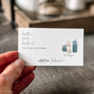 Può includere: Una carta bianca con il testo "breathe in, Behold, breathe out, Your King comes to you." e "adapted from Zechariah 9:9". La carta presenta un'illustrazione ad acquerello di tre candele nei toni del blu e del crema.