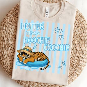 May include: A beige t-shirt featuring a graphic of a tiger wearing sunglasses and a straw hat while floating on a blue inner tube. The design includes light blue vertical stripes, star icons, and the text "HOTTER THAN A HOOCHIE COOCHIE".