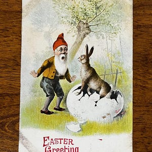 May include: Vintage Easter postcard featuring a surprised gnome and a rabbit emerging from a cracked egg. The scene is set in a garden with a tree and fence. The words "Easter Greeting" are printed in red.