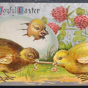 May include: A vintage Easter postcard featuring two chicks pecking at a worm. The postcard has a silver background with a large egg in the center and the text "A Joyful Easter" at the top.