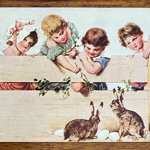 May include: Vintage Easter postcard featuring five children peering over a wooden fence. Two brown rabbits sit below, with several white eggs. The children are dressed in pastel colors, with one wearing a bonnet.