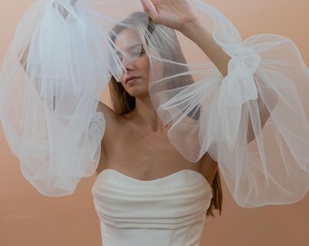 May include: A strapless white wedding dress with a fitted bodice and a flowing skirt. The dress is paired with sheer, voluminous sleeves and a veil, creating a romantic and ethereal look. The background is a soft peach color.