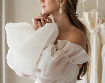 May include: A white off-the-shoulder wedding dress with a fitted bodice and a full skirt. The dress has large, puffy sleeves made of sheer fabric.