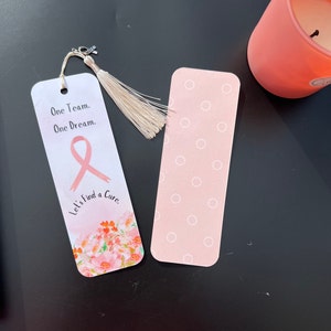 May include: Pink bookmark with a pink ribbon, flowers, and the text "One Team. One Dream. Let's Find a Cure."