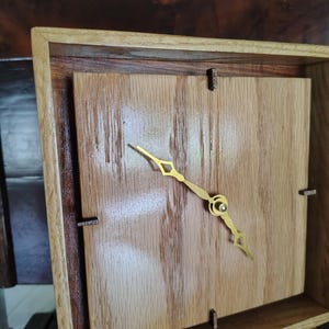 May include: A square wooden clock featuring a light oak face with a visible grain, set within a darker wood frame. The clock has gold-colored metal hands and small, dark wooden markers at the twelve, three, six, and nine o'clock positions.