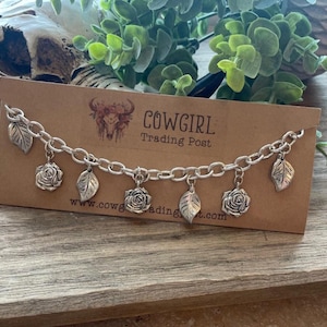 May include: Silver-toned charm bracelet featuring alternating rose and leaf charms. The bracelet is displayed on a brown card with the text "COWGIRL Trading Post" and a website address. The background includes greenery and a decorative skull.