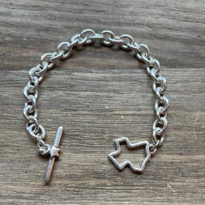 May include: Silver-toned bracelet featuring a chain link design. The bracelet includes a Texas-shaped charm and a bar and star clasp. The jewelry is displayed on a wooden surface.