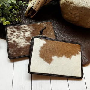 May include: Two rectangular zipper pouches crafted from cowhide featuring a natural brown and white spotted pattern. Each pouch is finished with a dark brown leather trim along the edges and includes a black zipper closure with a pull cord.