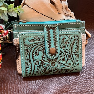 May include: A turquoise and brown leather wallet featuring an intricate floral embossed pattern, a snap button closure, and white stitching. The wallet includes sections of textured hair-on-hide leather and a metal zipper pull with a colorful fabric tassel.