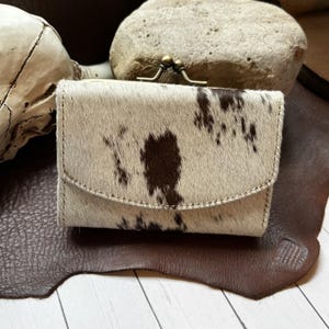 May include: A small coin purse featuring a cowhide pattern with white and dark brown fur. The purse has a curved flap closure and a metallic brass-toned kiss-lock clasp mechanism at the top, providing a secure way to store small items.