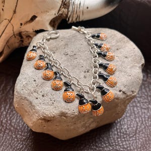 May include: A silver-toned chain link bracelet featuring multiple dangling charms. Each charm is an orange enamel jack-o'-lantern pumpkin wearing a black witch hat. The bracelet includes a toggle clasp closure and is constructed from metal links.