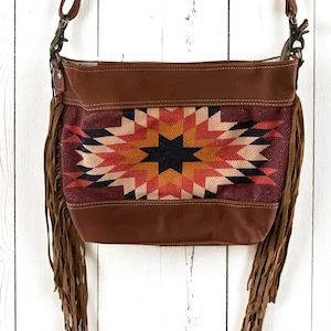 May include: Brown leather crossbody bag with a geometric patterned center in shades of red, orange, and black. The bag features long brown fringe and a brown leather strap. The bag is designed with a Southwestern theme.
