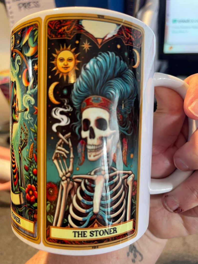 May include: A white ceramic mug featuring a colorful tarot card illustration of a skeleton with blue hair, wearing a headband and feathers, smoking a cigarette. The design includes a sun, moon, and stars, with the text THE STONER at the bottom.