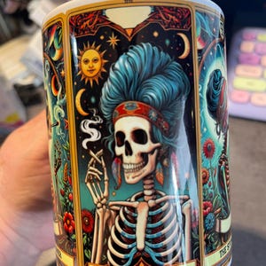 May include: A white ceramic mug featuring a colorful tarot card illustration of a skeleton with blue hair, wearing a headband and smoking. The design includes celestial elements like a sun and moon, floral accents, and the text THE STONER.
