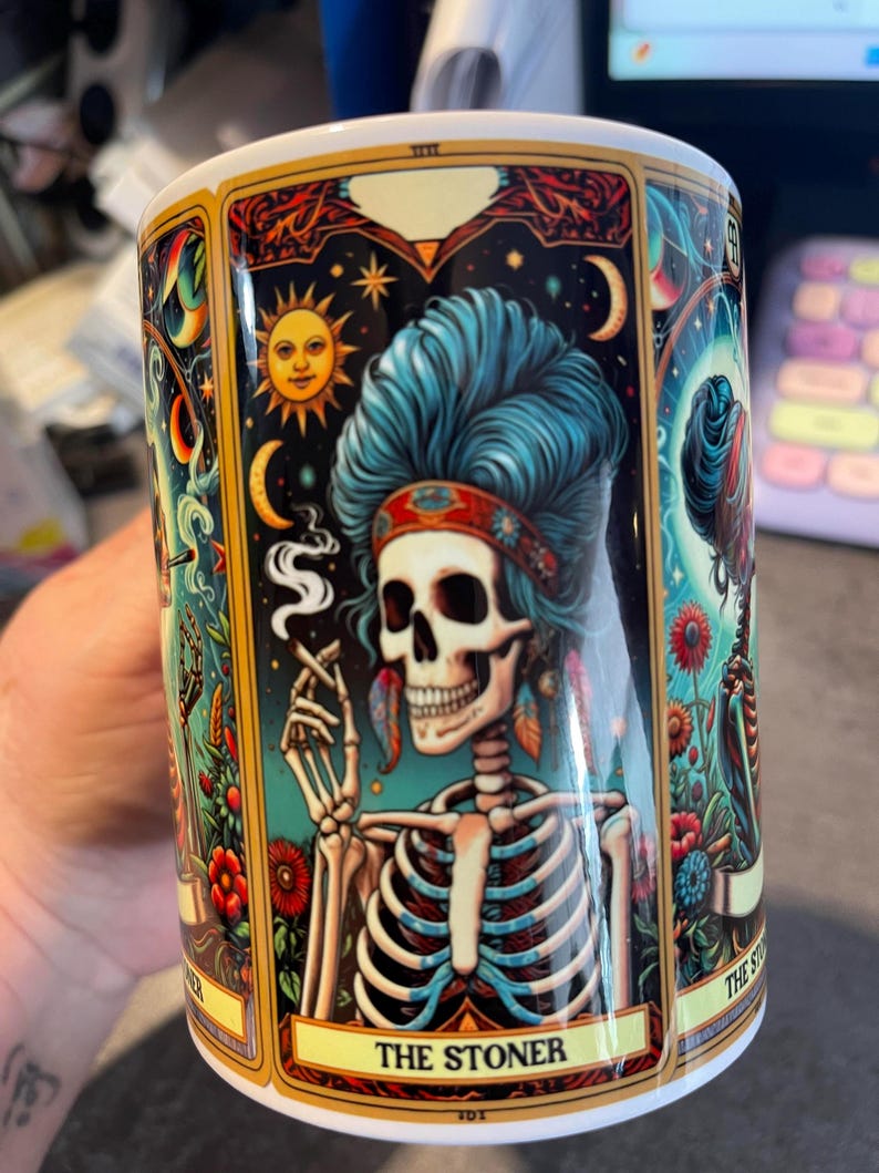 May include: A white ceramic mug featuring a colorful tarot card illustration of a skeleton with blue hair, wearing a headband and smoking. The design includes celestial elements like a sun and moon, floral accents, and the text THE STONER.