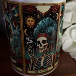 May include: A white ceramic mug featuring a colorful tarot card illustration of a skeleton with blue hair, wearing a headband and feathers, smoking a cigarette. The design includes a sun, moon, and stars, with the text THE STONER at the bottom.