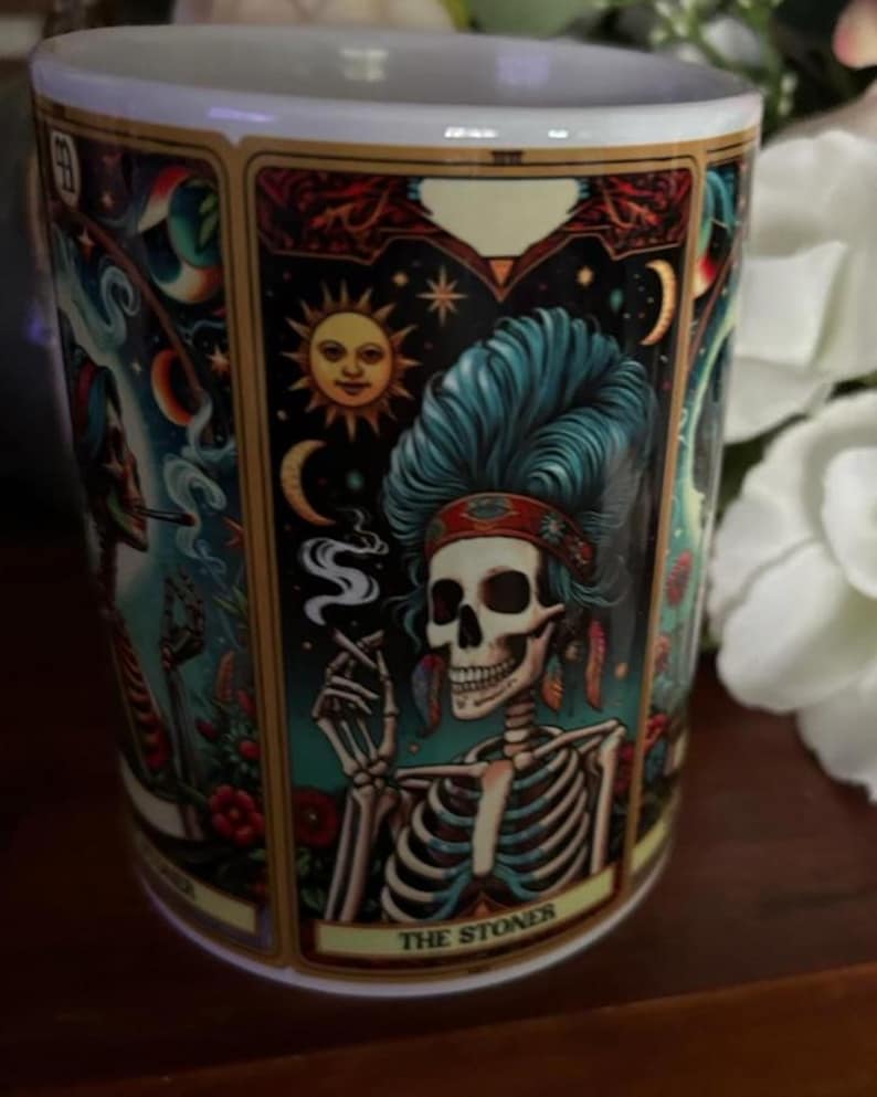 May include: A white ceramic mug featuring a colorful tarot card illustration of a skeleton with blue hair, wearing a headband and feathers, smoking a cigarette. The design includes a sun, moon, and stars, with the text THE STONER at the bottom.