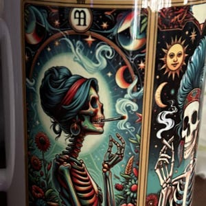 May include: A ceramic mug featuring a colorful illustration of two skeletons with hair, smoking, surrounded by celestial elements like a sun, moon, and stars, along with floral motifs. The design includes a central emblem with the letters AA.