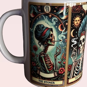 May include: A white ceramic mug featuring a colorful tarot card illustration of a skeleton with a bun hairstyle smoking a cigarette, surrounded by flowers and celestial symbols, with the text THE STONER printed at the bottom.