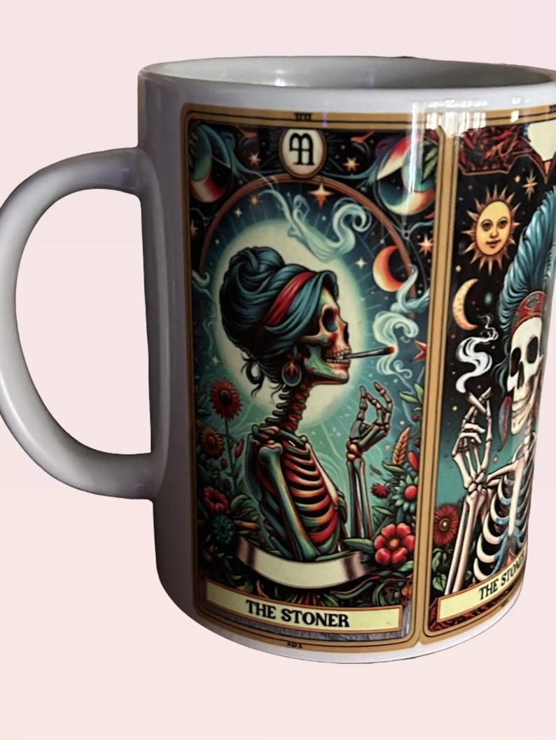 May include: A white ceramic mug featuring a colorful tarot card illustration of a skeleton with a bun hairstyle smoking a cigarette, surrounded by flowers and celestial symbols, with the text THE STONER printed at the bottom.