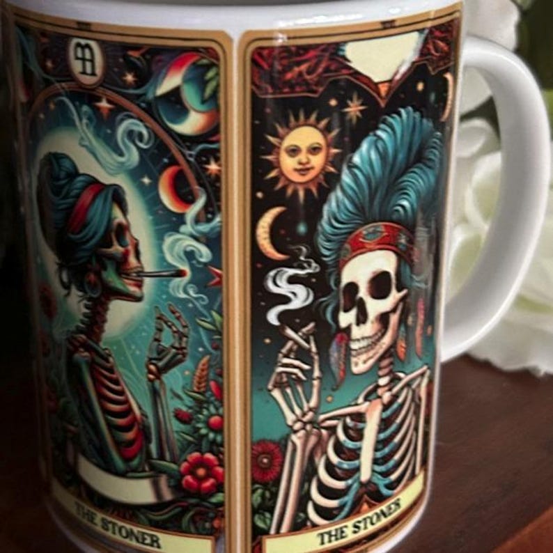May include: A white ceramic mug featuring two tarot-style illustrations of skeletons smoking. Each panel depicts a skeleton with colorful headwear, surrounded by celestial elements, flowers, and smoke, with the text THE STONER printed at the bottom.