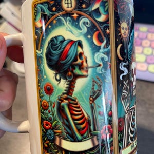 May include: A white ceramic mug featuring a colorful tarot card illustration of a skeleton with a red headband smoking a cigarette. The design includes vibrant flowers, a sun, and the text THE STONER at the bottom in a gold-bordered frame.