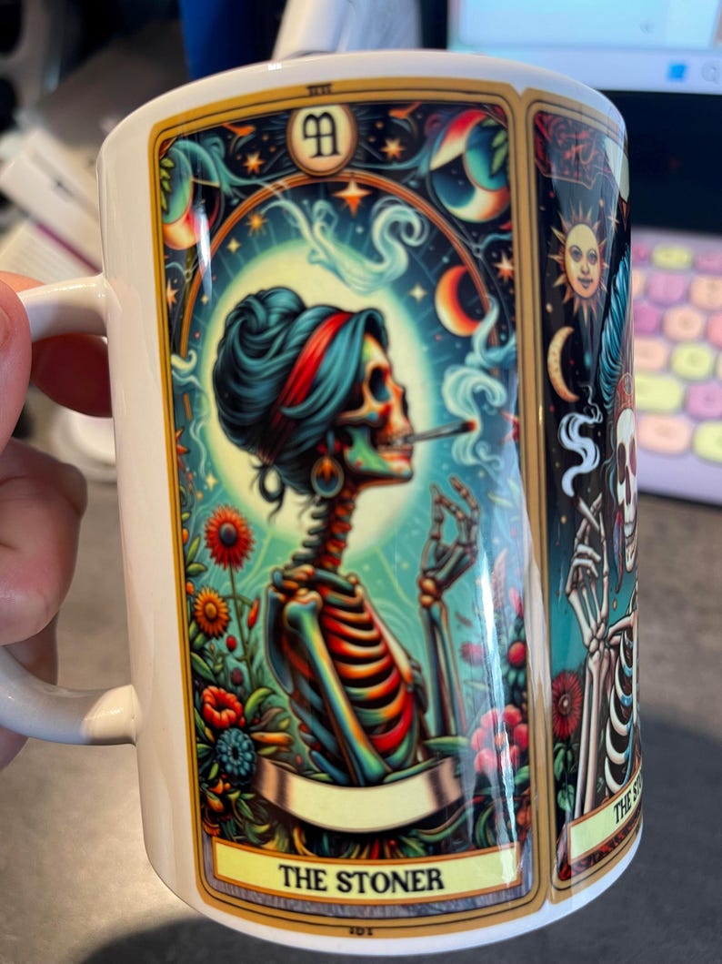 May include: A white ceramic mug featuring a colorful tarot card illustration of a skeleton with a red headband smoking a cigarette. The design includes vibrant flowers, a sun, and the text THE STONER at the bottom in a gold-bordered frame.