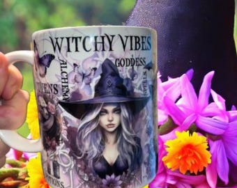 May include: A white ceramic mug featuring a graphic print of a person wearing a witch hat, surrounded by butterflies, flowers, and the text "WITCHY VIBES," "GODDESS," "ALCHEMY," "COVENS," "SPELLS," and "POTIONS" in a black serif font.