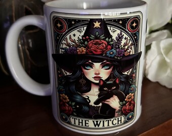 May include: A white ceramic mug featuring a colorful illustration of a person wearing a wide-brimmed hat adorned with flowers, holding a black cat. The design is framed in a decorative border with the text THE WITCH printed at the bottom.