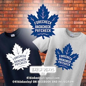 May include: Navy blue and light gray t-shirts featuring a white maple leaf design with the text "FORECHECK BACKCHECK PAYCHECK." A blue maple leaf graphic with the same text is also displayed. The image includes the text "INK N VINYL 416" and contact information.
