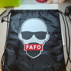 May include: Black drawstring bag with a white graphic of a person wearing sunglasses and a red rectangle with the text "FAFO" across the mouth.