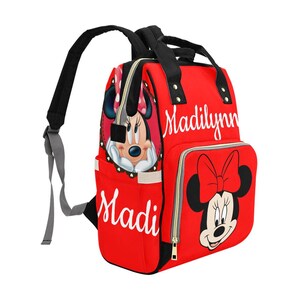 May include: Red backpack featuring Minnie Mouse graphics and the names "Madi" and "Madilynn" in white script. The bag has black straps and a zipper closure. A large Minnie Mouse face is on the front pocket.
