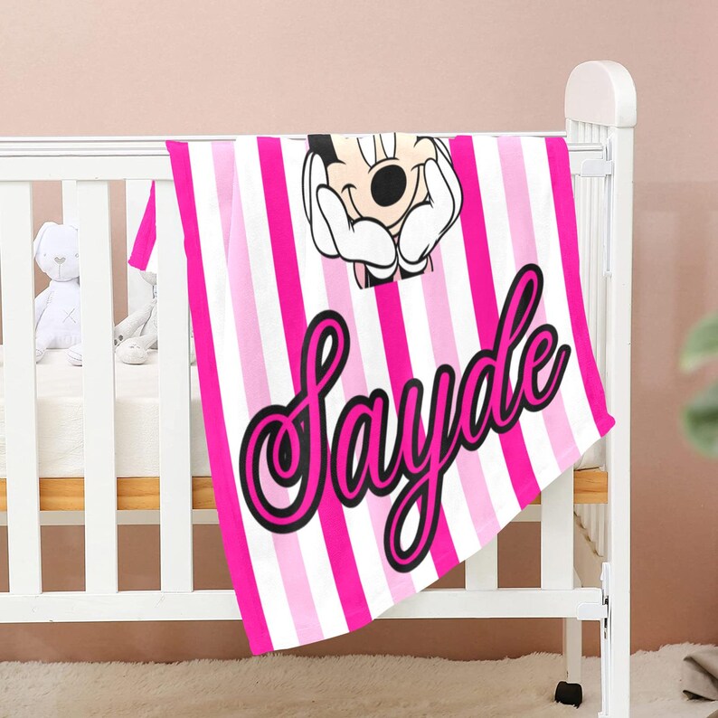 May include: A pink and white striped baby blanket with the name "Sayde" in a cursive font. The blanket features a cartoon image of a character's face. The blanket is draped over a white crib.