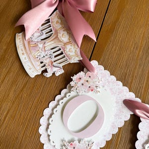 May include: A decorative banner featuring a carousel illustration with a dusty pink satin bow, paired with a scalloped cardstock cutout displaying the letter O, accented by soft pink floral illustrations and a delicate pink patterned background.