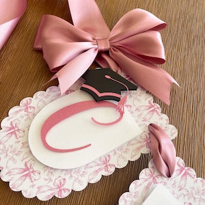 May include: Pink graduation banner with a pink satin bow and a black graduation cap. The banner is decorated with a white background with pink bows and flowers. The banner says "Class of 2023".