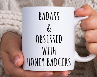 May include: White ceramic mug with the text "BADASS & OBSESSED WITH HONEY BADGERS" in black. The mug is held by a person wearing a cream-colored cable-knit sweater.