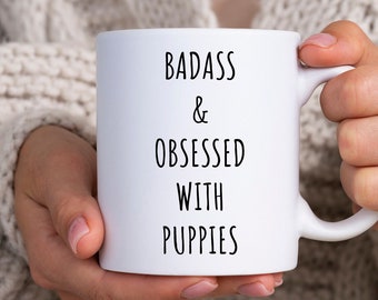 May include: White ceramic coffee mug with black text that reads "Badass & Obsessed With Puppies".