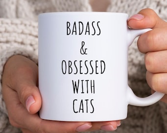 May include: White ceramic mug with black text that reads "Badass & Obsessed with Cats".