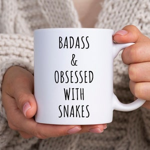 May include: White ceramic mug with black text that reads "Badass & Obsessed with Snakes".